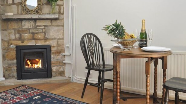 A cozy room with a stone fireplace, a round wooden table with chairs, a fruit bowl, and a bottle of champagne. - 1 Bed in Gloucester (oc-oldbn) (Upton Saint Leonards)