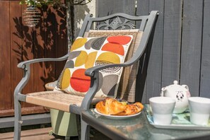 A wicker chair with a cushion featuring a geometric pattern, a table with a plate of food and teacups, and a hanging light fixture. - 2 Bed in Bideford (oc-swabr) (Higher Clovelly)