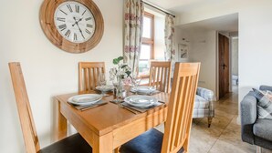 A dining area with a wooden table set for four, a clock on the wall, and a view into the living room. - 2 Bed in Bideford (oc-swabr) (Higher Clovelly)