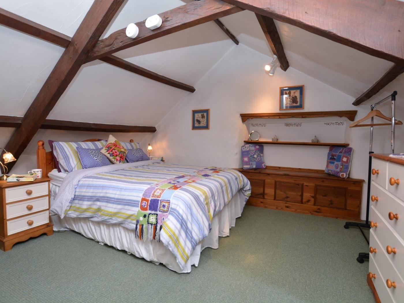 Une chambre confortable située sous les toits, avec un plafond en pente, un lit en bois recouvert d’une courtepointe à rayures, une table de chevet avec une lampe et une commode avec des tiroirs.