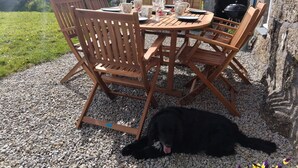 A round wooden table with chairs, a dog lying on the ground, and a stone wall in the background. - 4 Bed in Penzance (oc-holba) (Saint Buryan)