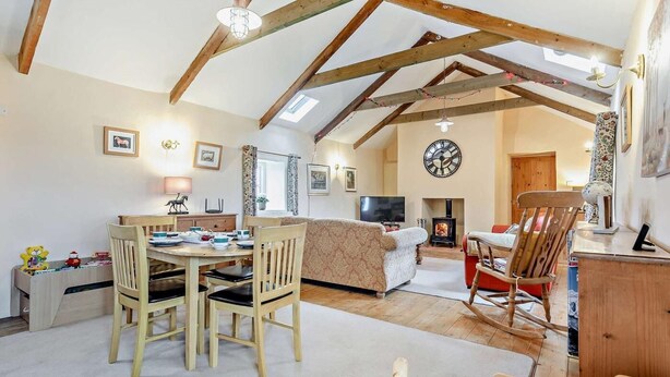 A cozy living room with a dining area, wooden beams, a fireplace, and a large clock on the wall. - 4 Bed in Penzance (oc-holba) (Saint Buryan)