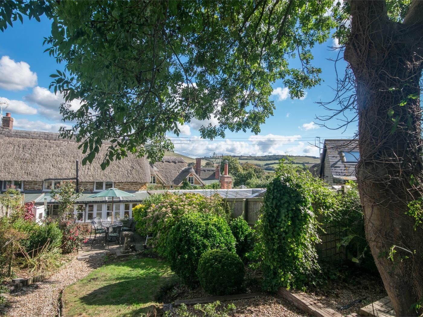 A traditional thatched-roof building with a garden area, surrounded by greenery and a clear sky.