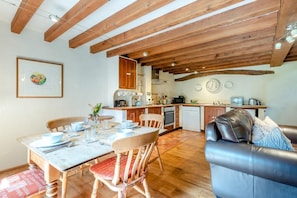 Ein gemütliches Wohnzimmer mit Ledersofa, Esstisch aus Holz und Stühlen. Im Hintergrund ist eine Küche mit Holzbalken und modernen Geräten zu sehen.
