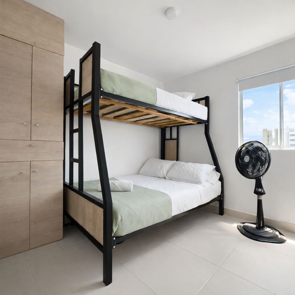 Uma cama beliche com estrutura de madeira, colchão verde e branco, e um ventilador preto no chão.