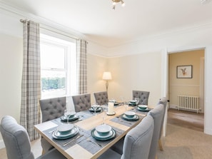 A dining room with a rectangular table set for six, upholstered chairs, a lamp, and a view of a garden through a window. - 3 Bed in Elgin (oc-ca294) (Elgin)