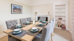 Un comedor con una mesa larga dispuesta para cuatro comensales, sillas grises tapizadas, una chimenea y una estantería con libros y objetos decorativos.