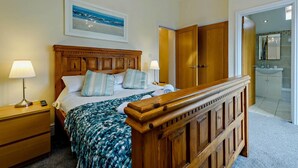 A bedroom with a bed, bedside lamps, a wooden headboard, a painting, and a bathroom visible through an open door. - 2 Bed in Tenby (oc-fb116) (Tenby)