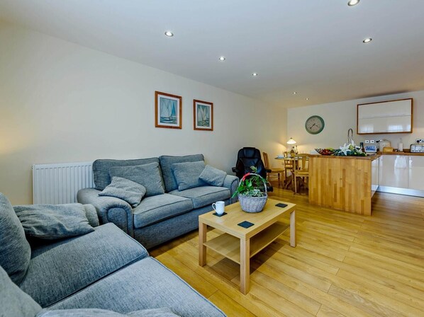A modern living room with a grey sofa, a wooden coffee table, and a dining area with a wooden table and chairs. - 2 Bed in Tenby (oc-fb116) (Tenby)