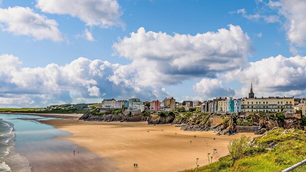 A coastal town with a sandy beach, buildings along the shore, and a prominent church spire. - 2 Bed in Tenby (oc-fb116) (Tenby)