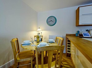 A dining area with a wooden table set for four, a wall clock, and a laptop on a counter. - 2 Bed in Tenby (oc-fb116) (Tenby)