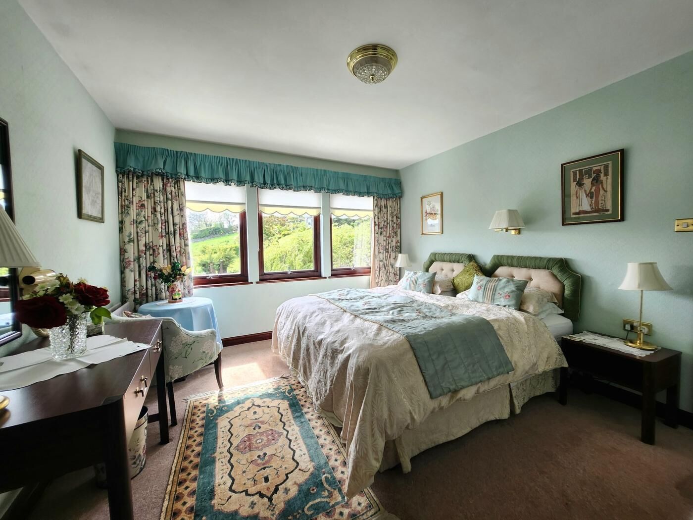 Une chambre à coucher comprenant un lit, des tables de chevet, un bureau, une chaise, une fenêtre avec des rideaux et une vue sur la verdure à l’extérieur.