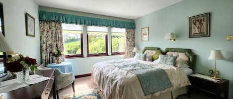 Une chambre à coucher comprenant un lit, des tables de chevet, un bureau, une chaise, une fenêtre avec des rideaux et une vue sur la verdure à l’extérieur.