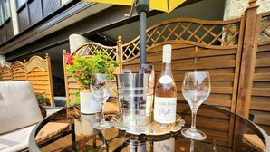 Een ronde glazen tafel met een wijnfles, twee wijnglazen, een metalen ijskoeler en een gele paraplu.