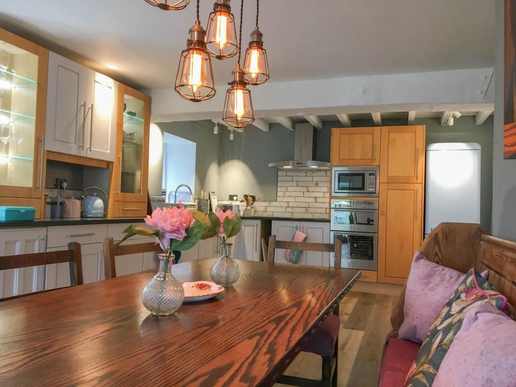 Une cuisine avec des meubles de rangement en bois, une table à manger agrémentée d’un vase de fleurs et des luminaires suspendus.