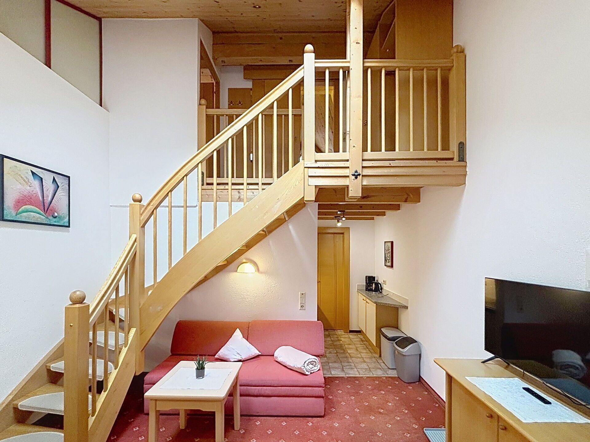 Ein Mehrstöckraum mit einer Holztreppe, einer Sitzecke, einem kleinen Tisch, einem Fernseher und einer Küchenzeile.