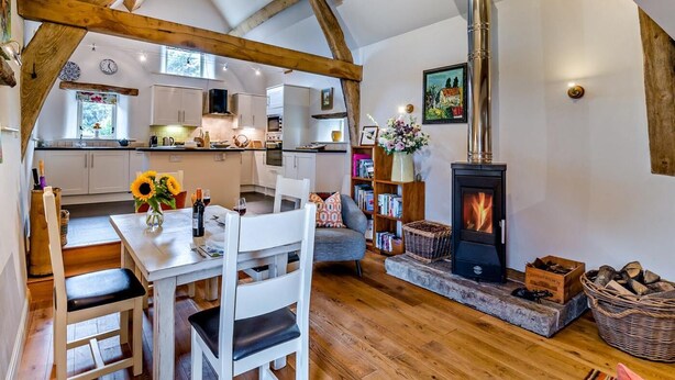 Un interior acogedor con vigas de madera a la vista, una zona de comedor con mesa y sillas, una chimenea y una cocina con armarios blancos y una ventana.