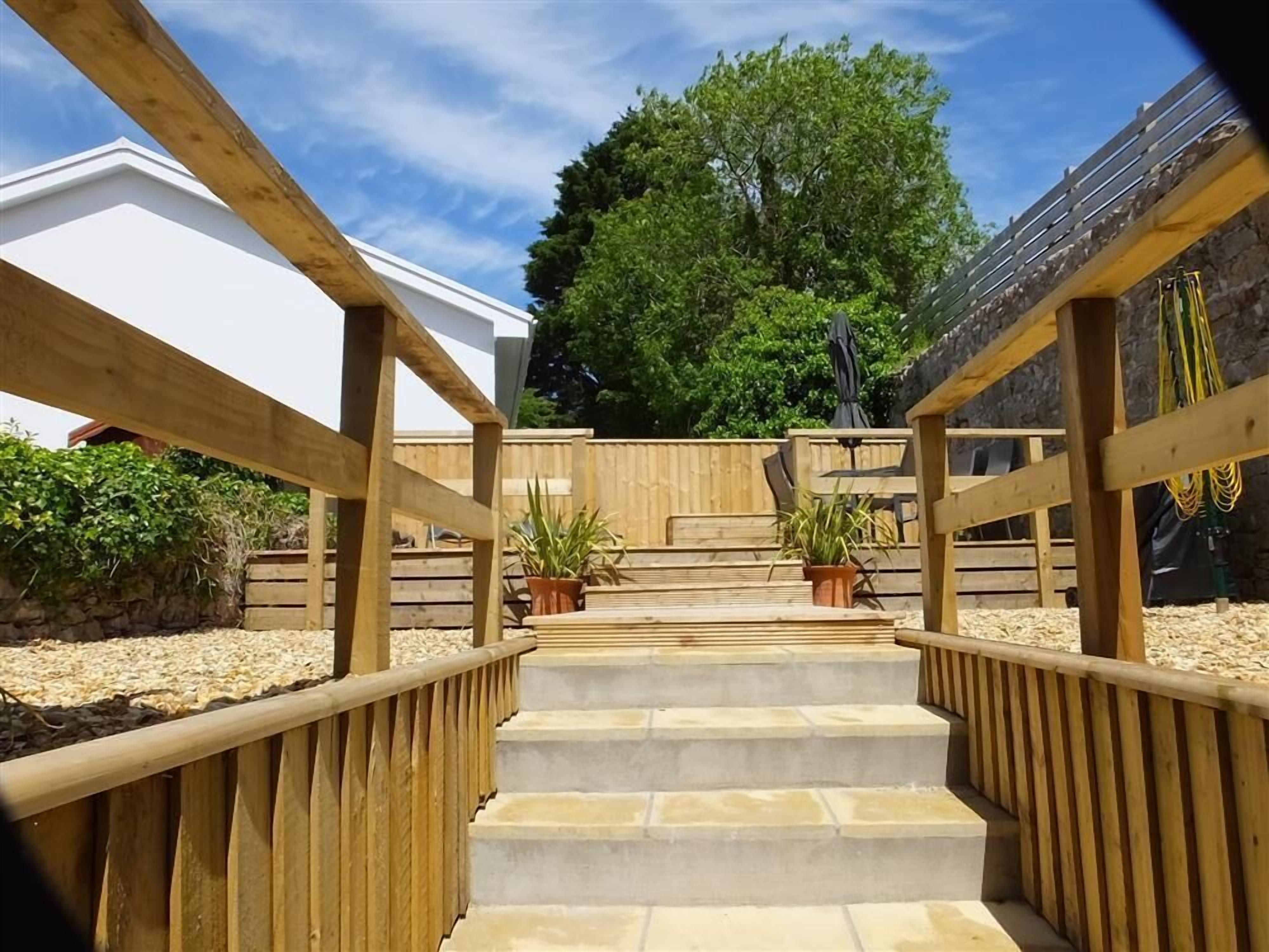 Una escalera de madera conduce a un espacio exterior cubierto con piso de piedra, rodeado de barandillas de madera y macetas con plantas.
