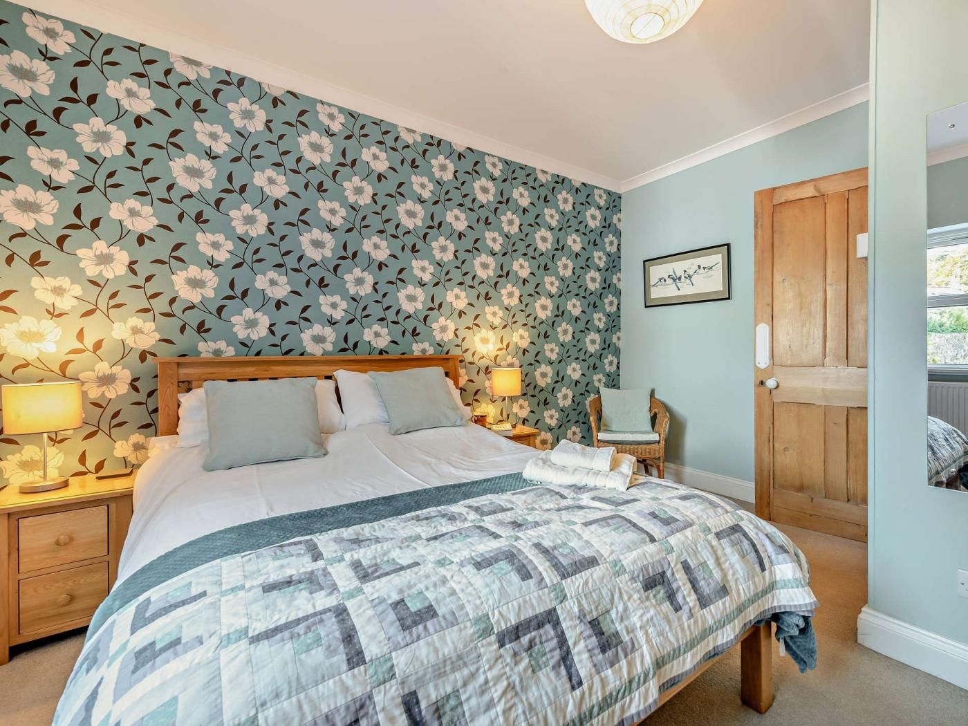 A bedroom with a floral wallpaper, a wooden bed frame, and a quilted bedspread.