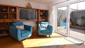 Un salon avec deux fauteuils bleus, un téléviseur, des étagères en bois et une porte-fenêtre coulissante donnant sur un jardin.