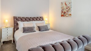 Une chambre à coucher avec une tête de lit capitonnée, un grand lit avec de nombreux oreillers, des tables de chevet avec des lampes et une œuvre d’art florale accrochée au mur.