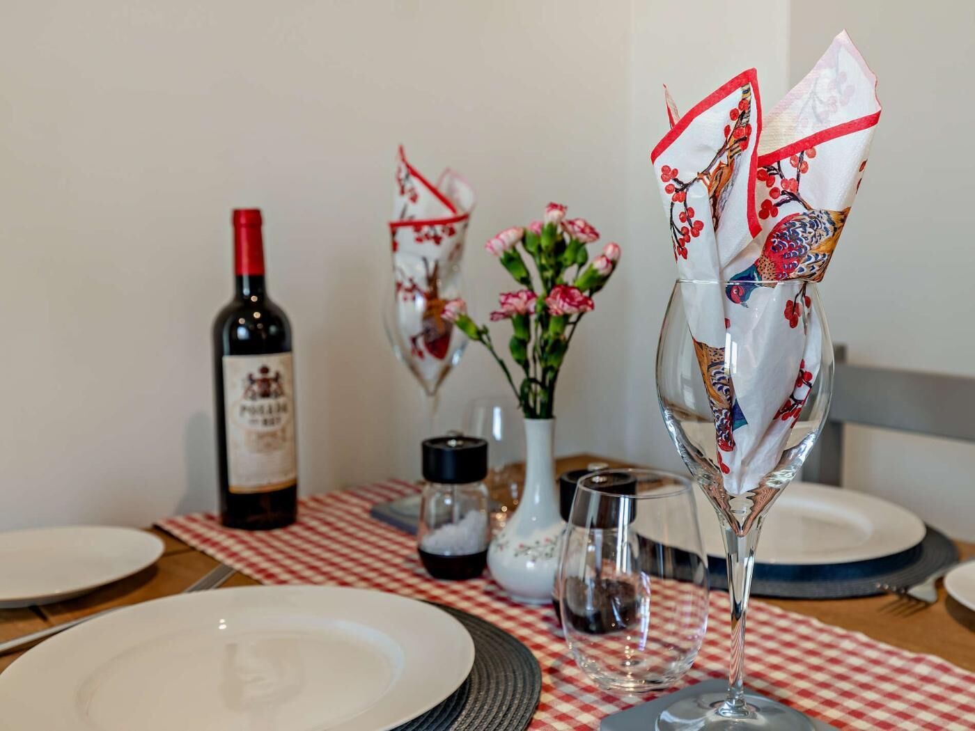 A dining table with a bottle of wine, a vase with flowers, and a glass with a decorative napkin.