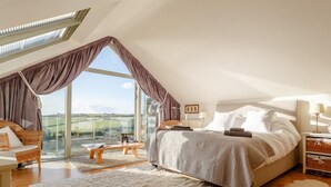 A bedroom with a large bed, a wooden bench, a fluffy rug, and a view of the countryside through a large window. - 4 Bed in Wembury (oc-54383) (Wembury)