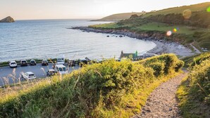 A coastal road leading to a parking area with cars and a small building, surrounded by greenery and a view of the sea. - 4 Bed in Wembury (oc-54383) (Wembury)