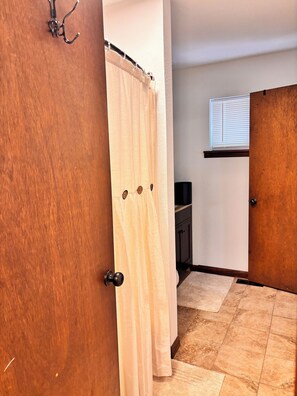 Quarto com porta de madeira, cortina branca, piso de azulejos, janela com persianas e um pequeno armário.