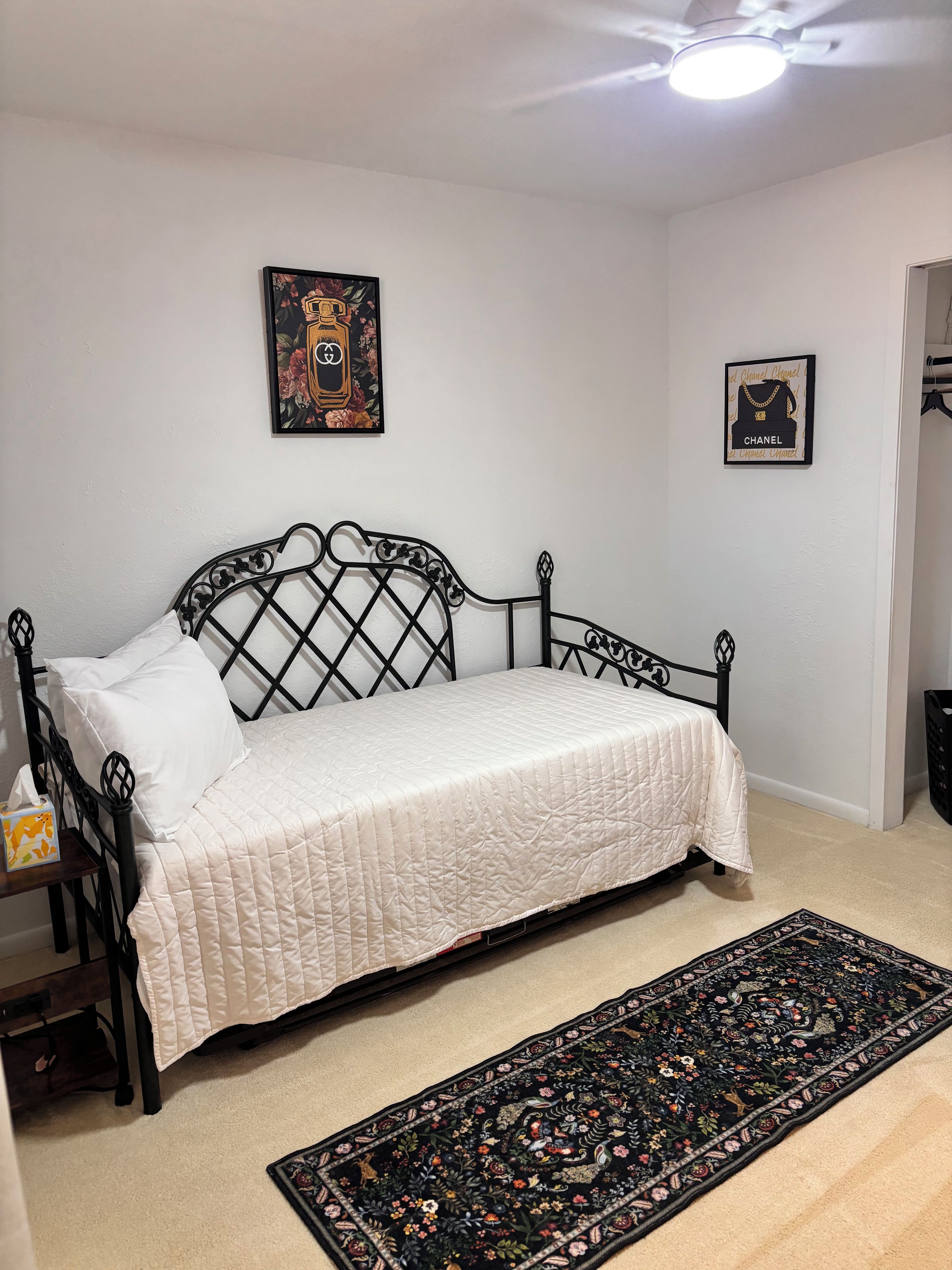 Ein Bett mit weißer Steppdecke, schwarzem Metallgestell, einem Blumenteppich und zwei gerahmten Bildern an der Wand.