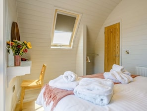 A bedroom with a sloped ceiling, a skylight, a wooden door, a bed with towels, a chair, and a shelf with flowers.