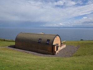A modern, curved wooden building with a small attached structure, situated on a grassy hill overlooking a large body of water.