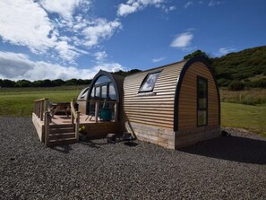 A small, wooden, semi-circular structure with a porch and a single window, set on a gravel surface with a grassy field and trees in the background.