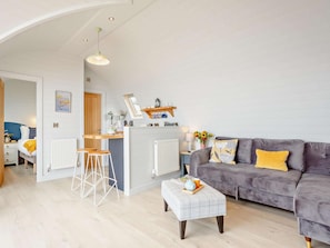 A modern living room with a grey sofa, a small white ottoman, and a kitchen area with a bar stool.