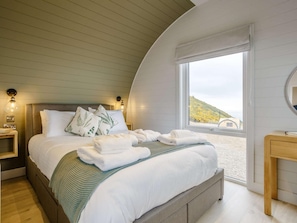 A bedroom with a curved ceiling, a large bed with white bedding, a bedside table with a lamp, and a view of a hillside through a window. - 2 Bed in Tain (oc-81340) (Nigg)