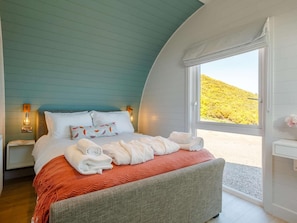 A bedroom with a curved ceiling, a bed with white linens, a view of a hillside, and a nightstand with a lamp.