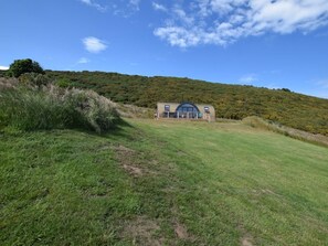 A single-story building with a large window and a door, surrounded by a grassy field and a hill in the background.