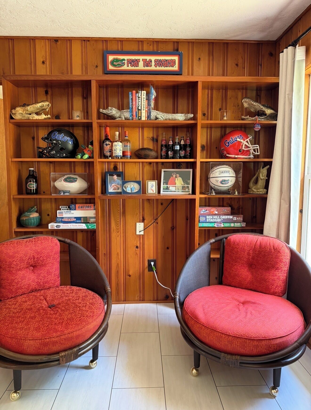 A room with two red cushioned chairs, wooden shelves with memorabilia, a sign reading 'Fear The Swamp,' and a window with curtains.