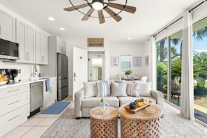 A modern kitchen with white cabinets, a stainless steel refrigerator, and a dining area with a sofa and coffee table. - Sandcastle Soul - Steps from Beach, Shared Pool, Shopping & Destin Attractions (Destin)