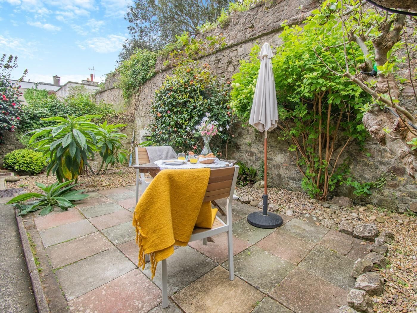 A patio with a table set for two, surrounded by greenery and a stone wall.