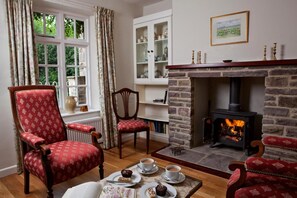 A cozy room with a fireplace, a red upholstered armchair, a wooden table with a book and a plate of food, and a window with curtains. - 3 Bed in Brecon (oc-bn161) (Talybont-on-Usk)