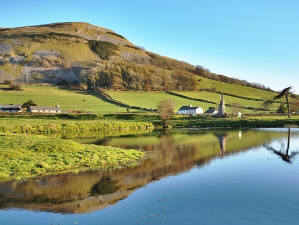 Ein ruhiger See mit der Spiegelung der umliegenden Landschaft, darunter ein Hügel und Gebäude.