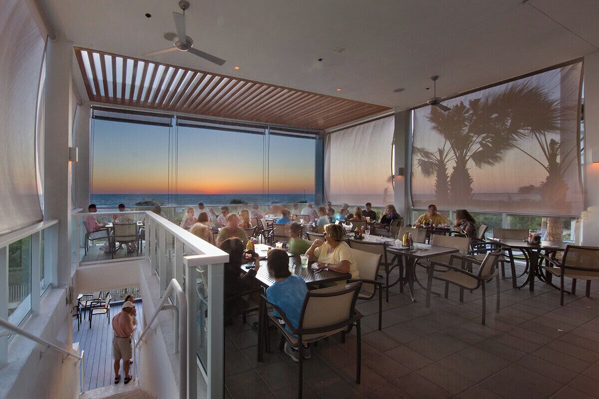 A dining area with tables and chairs, large windows with a view of the sunset, and a ceiling fan.