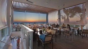 A dining area with tables and chairs, large windows with a view of the sunset, and a ceiling fan.