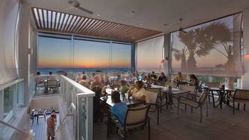 A dining area with tables and chairs, large windows with a view of the sunset, and a ceiling fan.