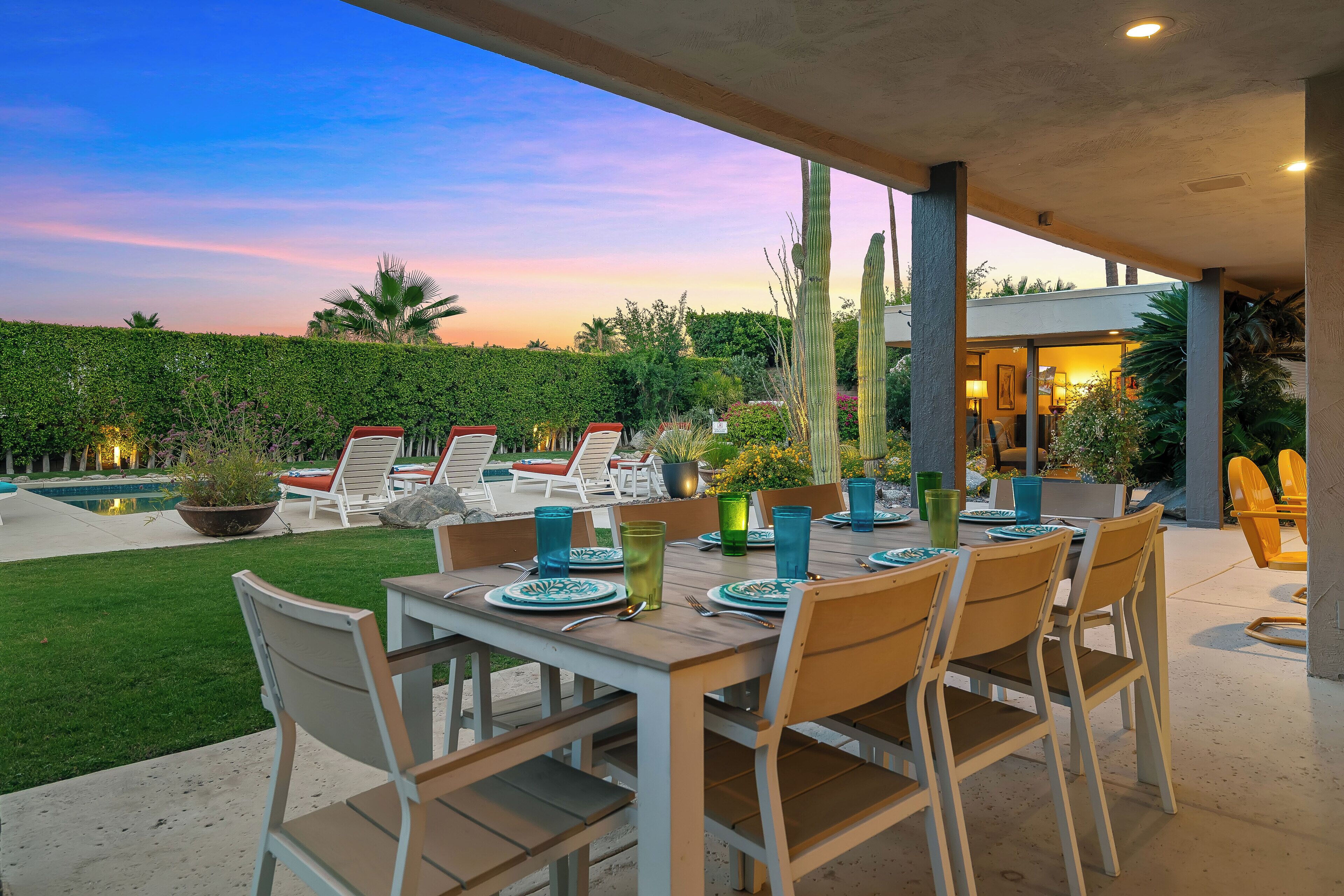An outdoor dining area with a table set for six, surrounded by chairs, and a pool in the background.