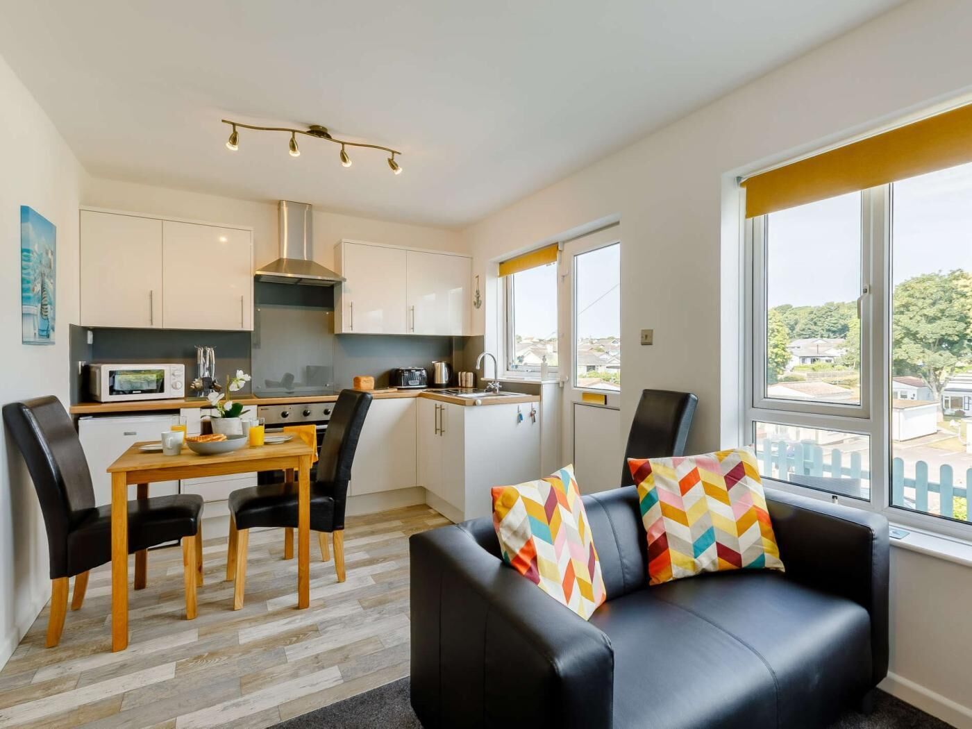 A modern kitchen with white cabinets, a dining table with chairs, a black leather sofa with colorful cushions, and large windows with a view of houses.