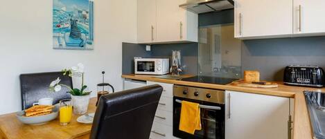 Una cocina compacta equipada con microondas, horno y tostadora. Incluye una mesa de comedor con una silla, una pequeña planta en maceta y un cuadro en la pared.