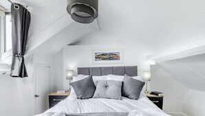 Una camera da letto moderna con soffitto spiovente, un letto con biancheria grigia, due lampade da comodino e un quadro incorniciato appeso al muro.