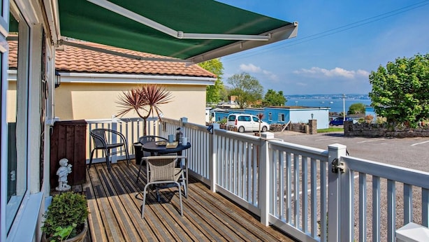 Eine Holzterrasse mit Tisch und Stühlen, ein grünes Markisenvorzelt und Blick aufs Meer.